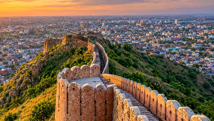 Ancient stone fort wall winding over green hills overlooking a sprawling city at sunset.